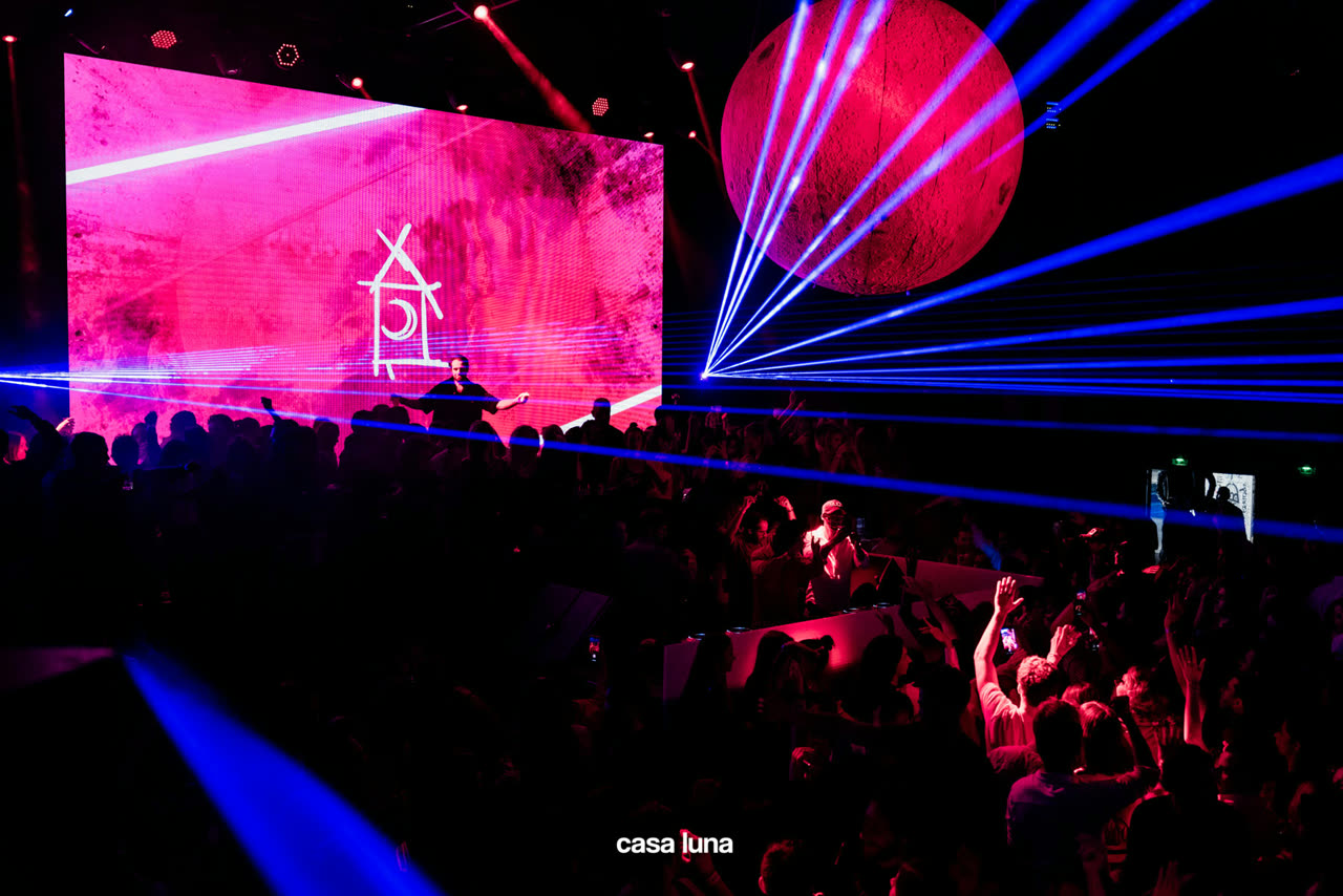 Casa Luna at Palais de Tokyo: a red moon hanging over the floor, blue lasers raking across a full room, the Casa Luna mark on the LED wall behind the booth.